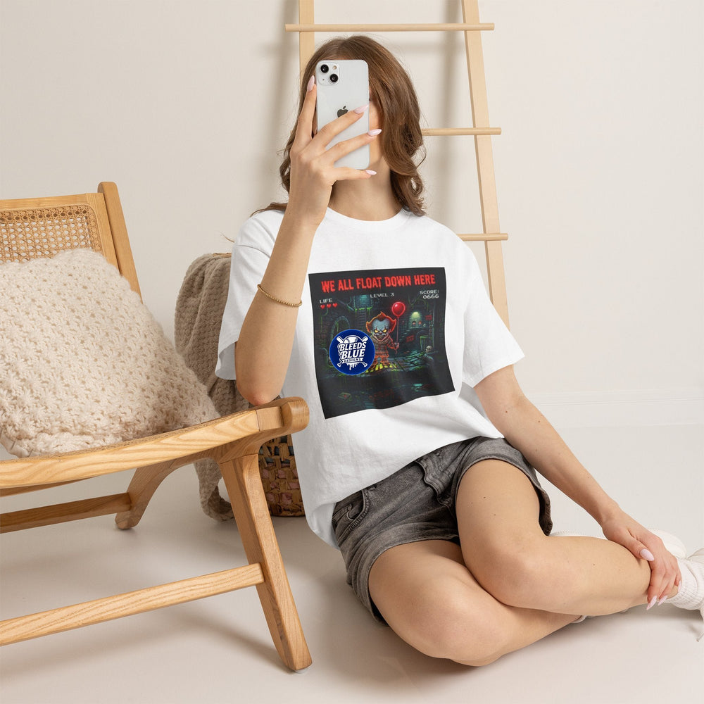 A woman takes a mirror selfie while wearing a white graphic t-shirt with a black and red 'Freddy Krueger's Dark Child' print, paired with gray denim shorts. She is sitting on a light-colored floor next to a wooden chair with a knit cushion and a wooden ladder against a neutral beige wall.