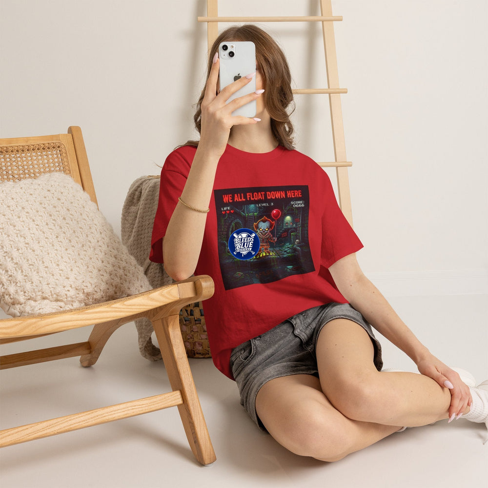 A woman takes a mirror selfie while wearing a red graphic t-shirt with a black 'ANCHORED.' print and gray denim shorts, sitting on a light-colored floor next to a wooden chair.